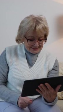 Yaşlı bir kadın rahat bir koltukta oturup tabletinde video görüşmesi yaparken gülümsüyor ve el sallıyor. Evde sanal bir sohbetten hoşlanıyor..