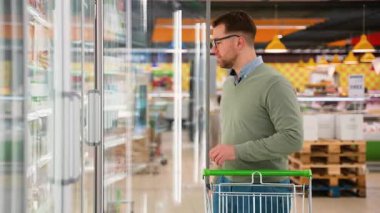 Yeşil kazaklı ve gözlüklü bir adam marketin soğutucu bölümünde indirimli yoğurda bakarken bir alışveriş arabasını itiyor..