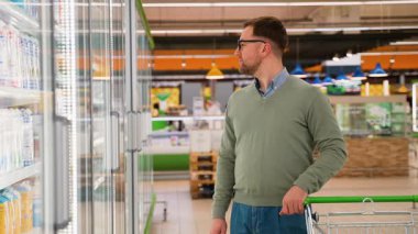Yeşil süveter ve gözlüklü bir adam, iyi aydınlatılmış bir marketin soğutulmuş kısmından süt seçerken bir alışveriş arabasını itiyor..