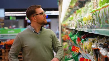 Yeşil kazaklı ve gözlüklü bir adam marketin sebze reyonunda taze salata seçeneklerine bakmış ve sağlıklı bir seçim yapmış..