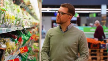 Yeşil kazaklı ve gözlüklü bir adam marketin sebze bölümünde paketlenmiş salata seçeneklerini inceleyerek sağlıklı bir yiyecek seçimi yapıyor..