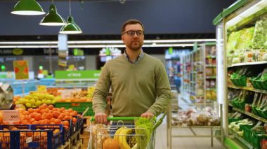 Yeşil kazaklı ve gözlüklü bir adam, marketin taze ürünler reyonuna bakarken muz, ananas, marul ve ekmekle dolu bir alışveriş arabasını itiyor..