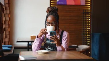 Gözlüklü genç bir Afro-Amerikalı kadın, bir kafede videolu sohbet için akıllı telefonunu kullanırken kahve içiyor.
