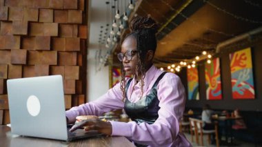 Gözlüklü Afrikalı Amerikalı bir kadın laptopunda görüntülü sohbet ederken şık bir kafede hamburger ve patates kızartması yiyor.