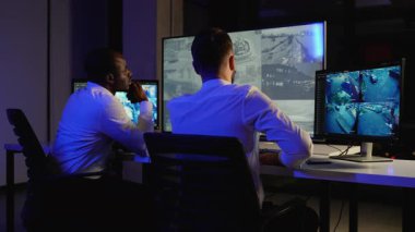 İki güvenlik görevlisi güvenlik, güvenlik ve risk yönetimi için güvenlik kamerası görüntülerini analiz ederek bir kontrol odasında birden fazla gözetleme monitörünü dikkatle gözlemliyor..