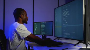 Bir yazılım geliştiricisi loş ışıklı bir ofiste çift monitörlerde programlama, siber güvenlik ve yazılım geliştirme üzerine çalışarak kod yazar ve hata ayıklar.