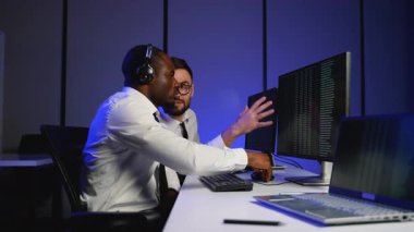 Bir veri merkezindeki iki bilişim uzmanı birden fazla monitörde gösterilen büyük verileri analiz edip tartışıyor. Veri yönetimi ve güvenliği konusunda işbirliği yapıyorlar..