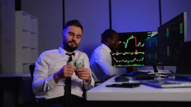 Takım elbiseli kendine güvenen bir tüccar borsa yatırımlarından elde ettiği kârı gösteren 100 dolarlık banknotları sayarken, meslektaşı da ekranlardaki mali tabloları izliyor..
