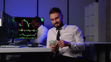Beyaz gömlekli gülümseyen bir tüccar dolarları sayarken, mali başarının keyfini çıkarırken, diğer tüccar birden fazla monitörde borsa grafiklerini analiz ediyor..