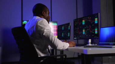 Beyaz gömlekli odaklı bir borsa simsarı birden fazla ekranda finansal grafikleri izler, pazar eğilimlerini analiz eder ve modern bir ofiste yatırım kararları alır..