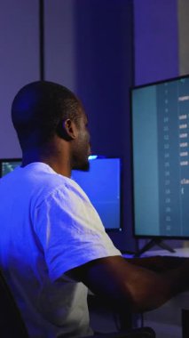 Odaklanmış bir Afrikalı Amerikalı yazılım geliştirici, programlama ve siber güvenlik üzerine çalışan loş ışıklı bir ofiste çift monitörlerde kod yazar ve hata ayıklar.