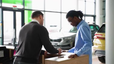 Bir araba galerisi çalışanı, arka planda arabalar olan modern bir galeride bir araç satın alma sözleşmesi imzalayan bir müşteriye yardımcı olur..