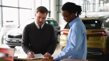 Bir araba satıcısı, galeride bir müşteriye, bir araç alımına ilişkin evrak işlerinde yardımcı olan kontrat detaylarını açıklar..