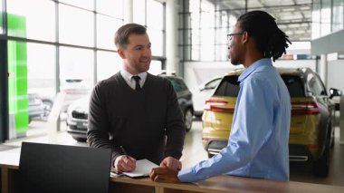 Bir araba satıcısı, modern bir galeri galerisinde memnun bir müşteriyle indirimli bir araç satışını sonuçlandırır ve satın alma anlaşmasını tartışır..