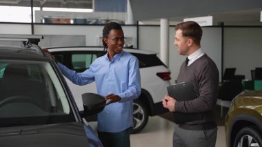 Gülümseyen bir müşteri, bir araba galerisinde bir aracı incelerken bir araba satıcısıyla konuşur, karar vermeden önce özellikleri ve satın alma seçeneklerini tartışır..