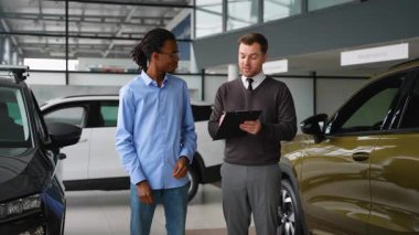 Bir araba satıcısı, yeni bir aracın avantajlarını bir galeri galerisinde potansiyel bir alıcıya, özellikleri, performansı ve fiyatlandırma seçeneklerini ön plana çıkararak açıklar..