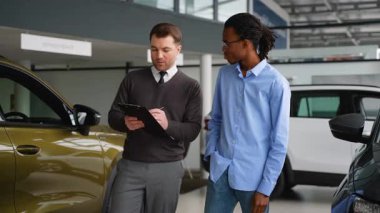 Bir araba satıcısı panoyu inceler ve satın almadan önce galerideki bir müşteriye yeni bir aracın özelliklerini ve fiyatlandırmasını açıklar..