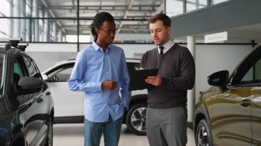 Bir araba satıcısı, Afro-Amerikalı bir müşteriye galeride araç seçmesinde yardımcı olur. Satın almadan önce özellikleri, fiyatları ve finansman seçeneklerini tartışır..