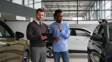 Bir araba satıcısı ve Afrikalı bir Amerikalı müşteri karar vermeden önce bir galeride araç seçeneklerini, özellikleri, fiyatları ve finansmanı gözden geçirirler..