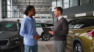 Bir araba satıcısı ve bir müşteri, başarılı bir araç alımını tamamladıktan sonra galeride el sıkışıyor ve alışverişi tatmin edici bir şekilde tamamlıyorlar..