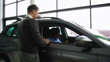 Bir araba satıcısı, bir galeride sağda oturan bir müşteriye satış yapmadan önce onun özelliklerini açıklayarak yardımcı olur..