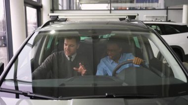 Bir araba satıcısı müşterinin yanında oturur ve satın almadan önce aracın özelliklerini açıklar. Alıcı direksiyonu modern bir araba galerisinde tutuyor..
