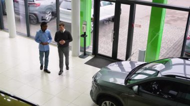 Bir araba satıcısı, müşterisiyle birlikte modern bir galeride araç seçeneklerini tartışıyor. Büyük cam pencereler dışarıda park halindeki arabaları gösteriyor..