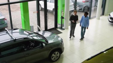 Bir araba satıcısı, müşterisiyle birlikte modern bir galeride araç seçeneklerini tartışıyor. Büyük cam pencereler dışarıda park halindeki arabaları gösteriyor..