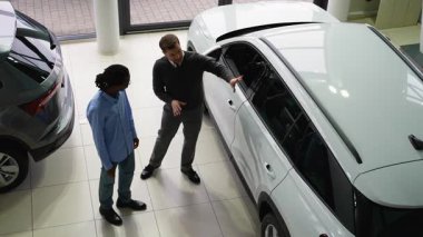 Bir araba satıcısı, satış mağazasında potansiyel bir alıcıya beyaz bir SUV sunar. Satın alma kararı için tasarımını ve özelliklerini vurgular..
