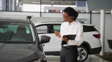 Beyaz tişörtlü profesyonel bir araba satıcısı galeride bir aracı inceler. Elinde bir not panosu ve kalemle arabaların detaylarını inceler..