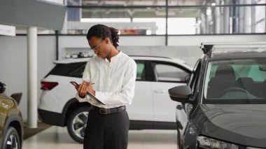 Beyaz gömlekli Afro-Amerikalı bir araba satıcısı araba galerisinde envanter çıkarıyor, satılık araçları incelerken panoya yazı yazıyor..