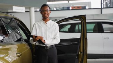 Beyaz gömlekli gülümseyen bir araba satıcısı, modern bir galeride bir aracı incelerken elinde bir pano tutuyor. Kaliteyi ve detayları sağlıyor..