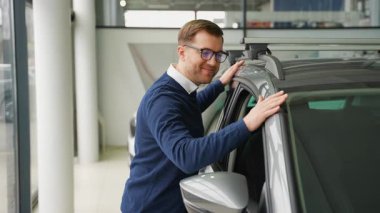 Gözlüklü gülümseyen adam modern bir galeride yeni bir arabayı teftiş ediyor, satın almadan önce araç tasarımı ve özelliklerini takdir ediyor..