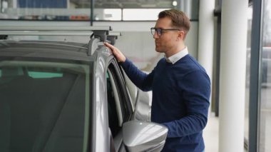 Gözlüklü gülümseyen adam galeride yeni aldığı arabaya hayran, yeni bir araca yaptığı yatırımdan memnun ve heyecanlı..