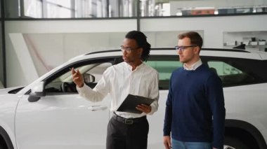 Profesyonel bir araba satıcısı, beyaz bir SUV 'nin özelliklerini modern bir galerideki potansiyel bir müşteriye açıklar..