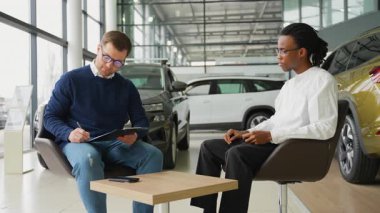 Bir müşteri araba satın alma anlaşmasını imzalarken, bir satıcı galeride gözlemler ve yeni bir araç için son kararı verir..