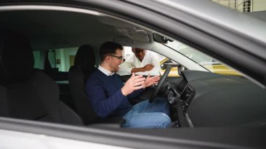 Bir araba satıcısı, modern bir galeride sürücü koltuğunda oturan potansiyel bir müşteriye aracın iç hatlarını açıklar..