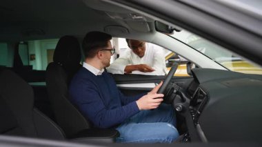 Bir araba satıcısı, bir satış şirketinde sürücü koltuğunda oturan bir müşteriye satın alma kararı vermesine yardımcı olan bir aracın özelliklerini açıklar..