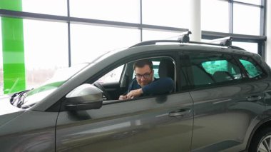 Neşeli bir adam yeni satın aldığı arabasında oturuyor, gülümsüyor ve anahtarları tutuyor, yeni arabası için heyecanlanıyor..