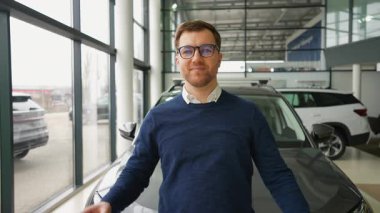 Neşeli bir adam araba galerisinde duruyor, yeni bir araba alırken gülümsüyor. Heyecanlı ve yeni arabasını kullanmaya hazır..