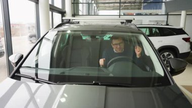 Neşeli bir adam, yeni aldığı arabasında bir galeride oturmuş, heyecanla kutluyor. Yumruklarını kaldırır ve mutluluk içinde dans eder..