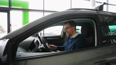 Neşeli bir adam, yeni aldığı arabanın şoför koltuğunda oturuyor. Yeni arabası için heyecandan ve mutluluktan gülümsüyor..