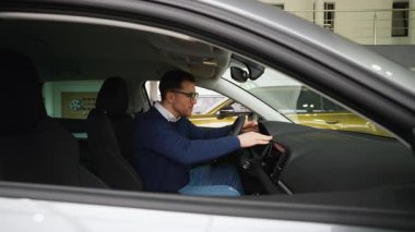 Neşeli bir adam yeni aldığı arabasında oturuyor, heyecanla satın alışını kutluyor. Gülümser ve mutlu bir şekilde hareket eder. Satıcılıktan zevk ve memnuniyet duyar..