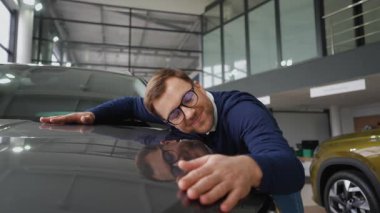 Keyifli bir adam yeni satın aldığı arabanın kaputuna sarılıyor, galeriden satın almaktan duyduğu haz ve memnuniyeti dile getiriyor..