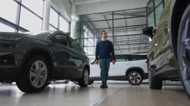 Şık bir adam parlak bir araba galerisinde yürüyor, çeşitli araçları keşfediyor ve bir galeriden yeni bir şey almak için seçenekleri değerlendiriyor..
