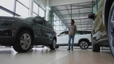 Genç bir adam parlak bir araba galerisine gider, çeşitli araçları inceler ve bir sonraki araba alımı için seçenekleri değerlendirir..