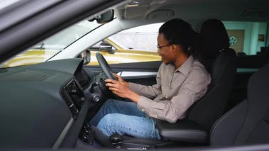 Afro-Amerikalı bir adam sağ el sürüş aracının sürücü koltuğunda oturur, direksiyonu dikkatle inceler ve satın almadan önce kontrol eder..