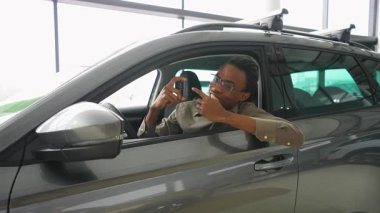 Neşeli bir Afrikalı Amerikalı adam yeni aldığı arabasında oturuyor, gülümsüyor ve gururla modern bir galerinin anahtarlarını gösteriyor..