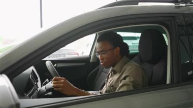Neşeli bir Afrikalı Amerikalı adam yeni satın aldığı arabasında oturuyor, direksiyonu tutuyor ve modern bir galeride gülümsüyor..