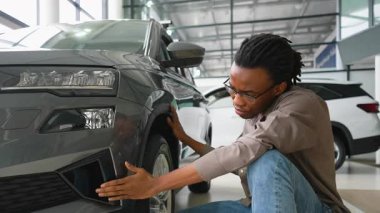 Odaklanmış Afro-Amerikalı bir adam, satın almadan önce modern bir galeride yeni bir arabanın dış ve arka tamponunu yakından inceliyor..